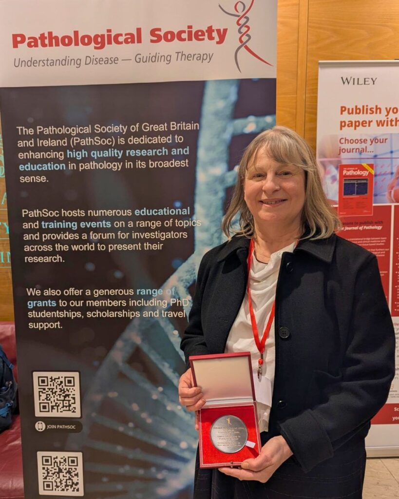 Professor Louise Jones stands smiling, holding a box containing the Goudie medal. She stands in front of a banner for the Pathological Society.