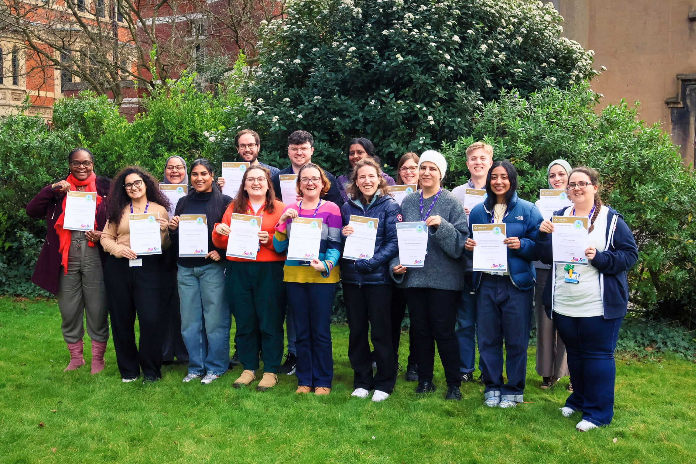 A group of BCI staff stand on the green, smiling at the viewer and holding up their LEAF bronze and silver certificates