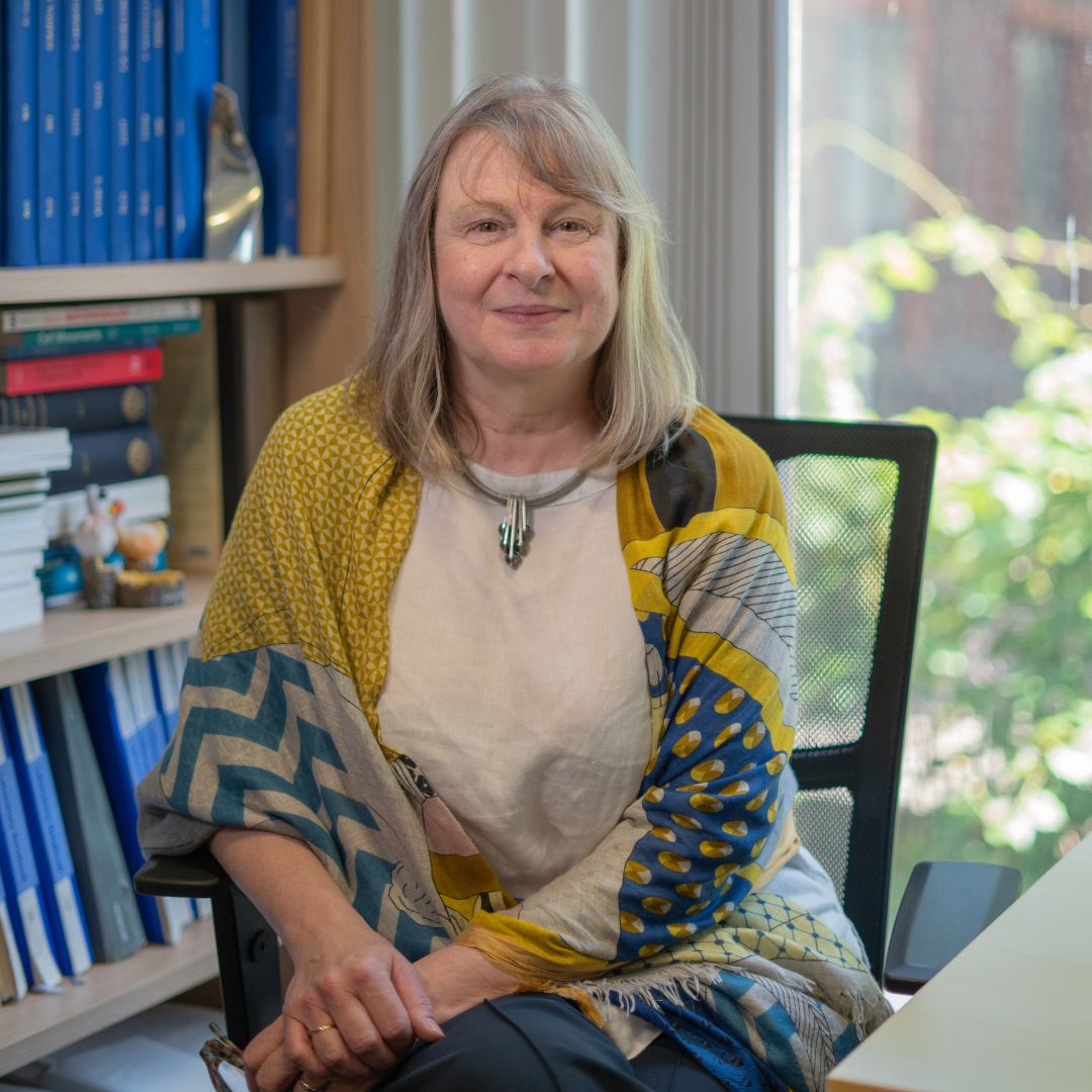 Professor Louise Jones sits in an office chair beside a desk, wearing a light top and a patterned shawl with yellow, blue, and grey designs. Behind them is a bookshelf filled with blue and red books and a few decorative items. A window in the background lets in natural light and shows greenery outside, creating a professional, tidy workspace.